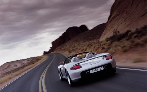 A sleek Porsche Carrera GT speeds along a winding mountain road, showcasing its dynamic design against a dramatic backdrop of rocky terrain and moody skies.