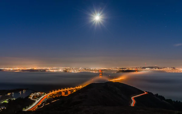 HD PC desktop wallpaper and background of the man-made Golden Gate Bridge at night — towers piercing fog, city lights and winding car light trails beneath a bright moon.