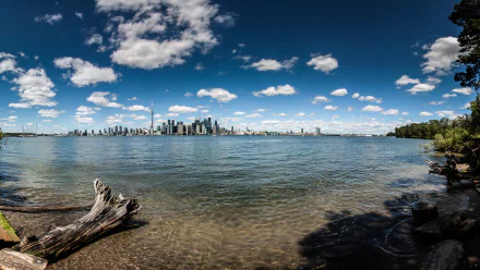 HD desktop wallpaper of Toronto’s man-made skyline across the water under a bright blue sky with scattered clouds, featuring natural shoreline elements in the foreground.