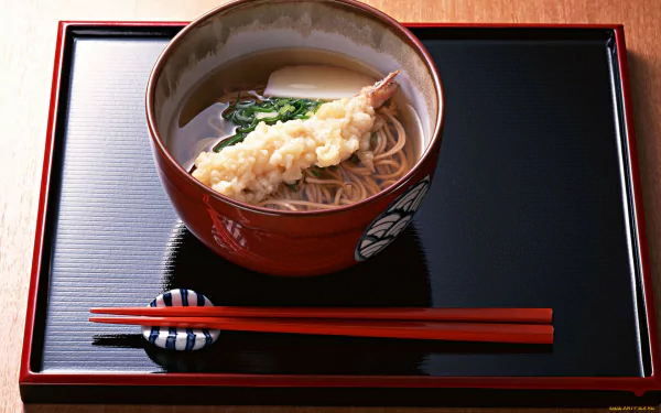 A vibrant bowl of Japanese soba noodles topped with tempura, garnished with vegetables, set on a sleek black tray with red chopsticks, serving as an appealing HD PC desktop wallpaper.