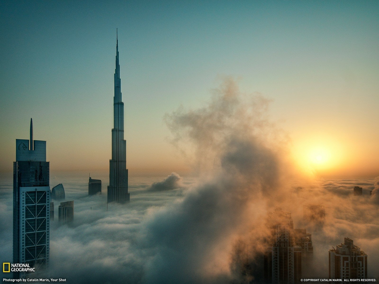 A stunning HD desktop wallpaper showcasing Dubai's skyline, featuring the Burj Khalifa rising above a sea of clouds at sunrise, with a warm glow illuminating the scene.