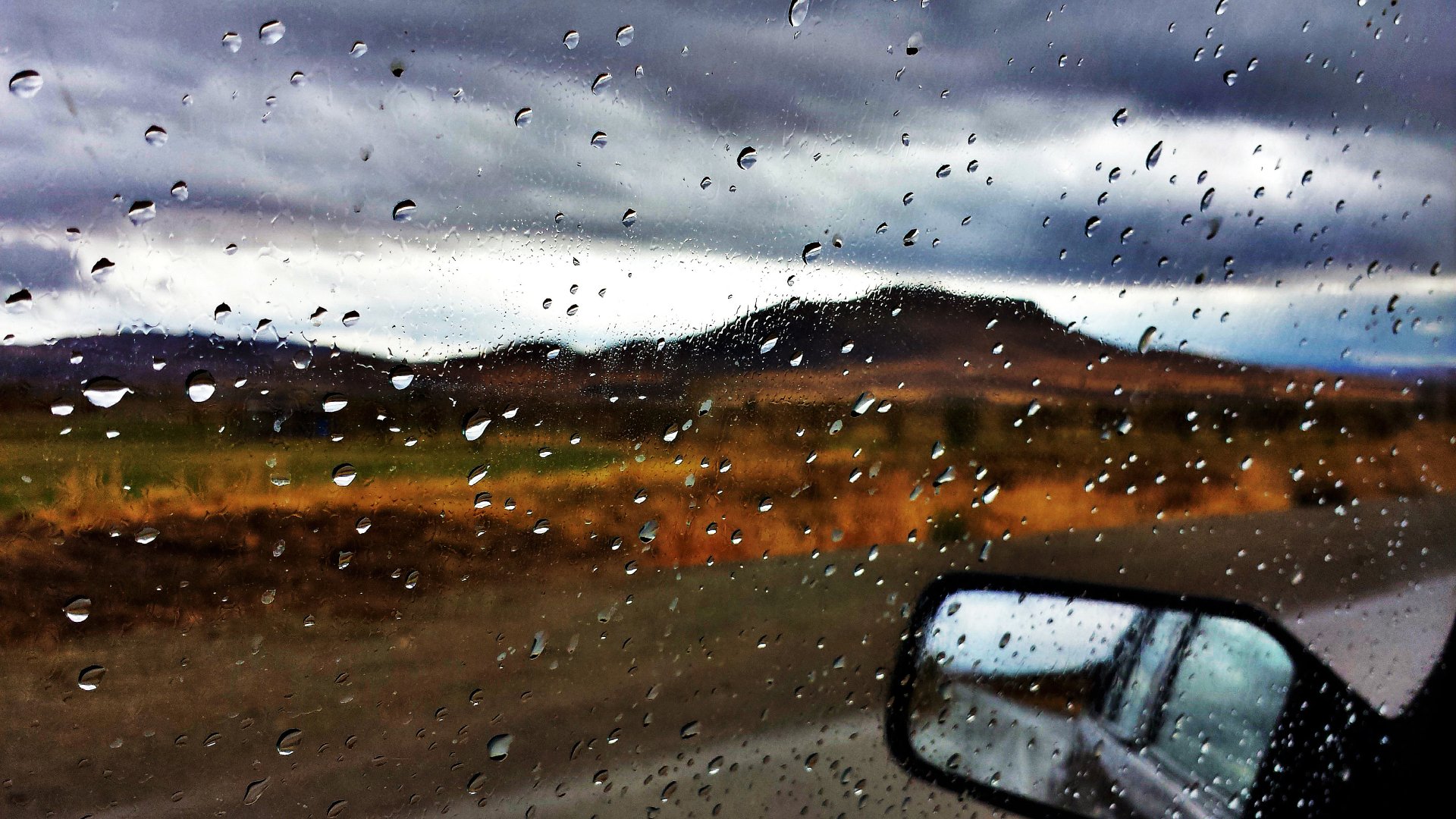A scenic view from a car window in Kordestan, Iran, showcasing water droplets on the glass, capturing the essence of fall with a dramatic sky and distant hills.