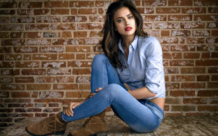 Brunette model Irina Shayk poses stylishly against a brick wall, wearing a light blue shirt and denim jeans, showcasing her bold lipstick in this HD wallpaper.