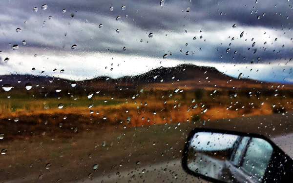 A scenic view from a car window in Kordestan, Iran, showcasing water droplets on the glass, capturing the essence of fall with a dramatic sky and distant hills.