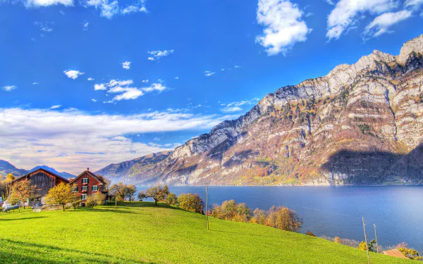 HD wallpaper of a tranquil riverside scene with green fields, a house, and a mountain range under a blue sky with scattered clouds.