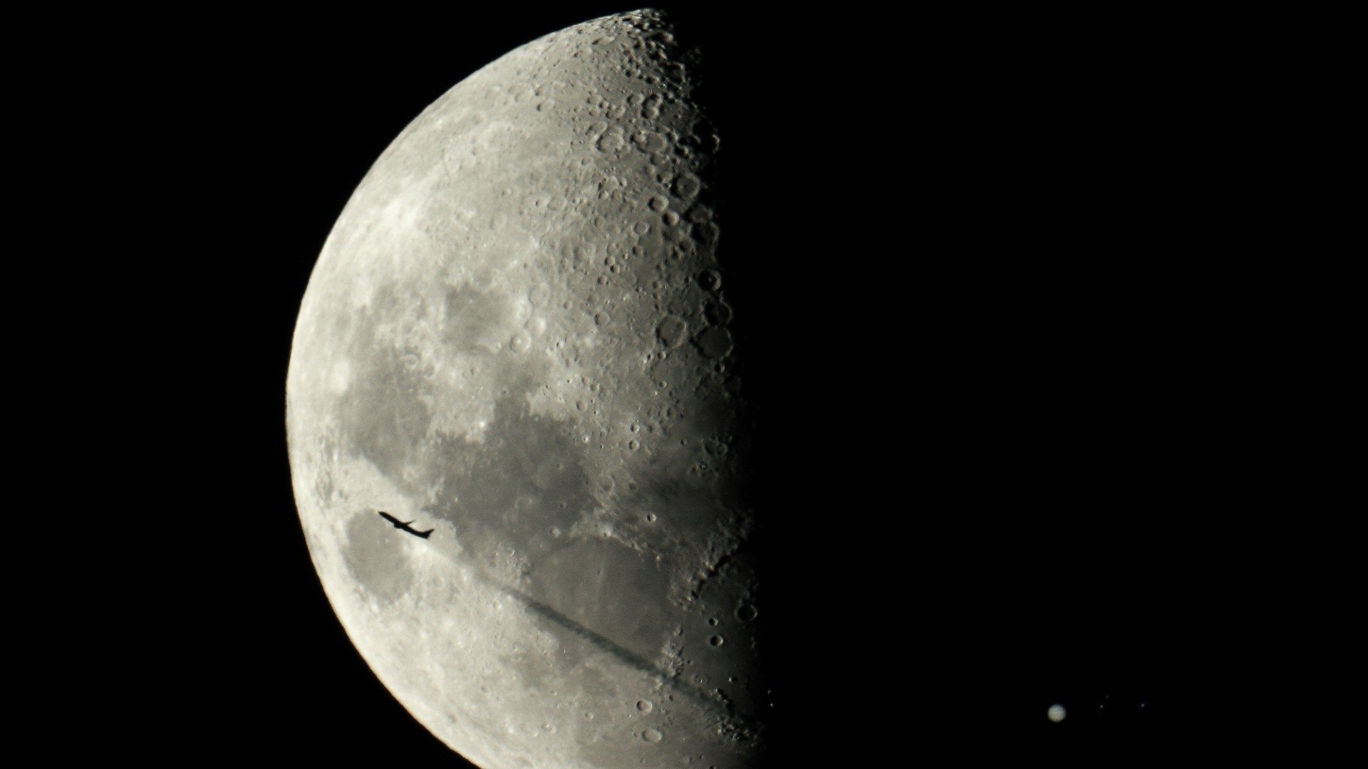 HD PC desktop wallpaper of nature's moon: close-up of the illuminated half against a black sky, a small silhouetted object crosses the lunar disk with two star-like points nearby.