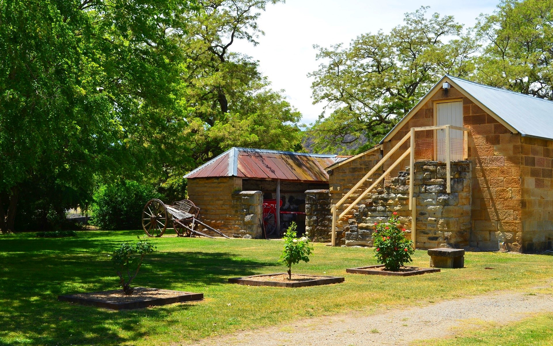 A garden scene at Eskbank House in Lithgow featuring a rustic stone building, an old cart, and lush green trees under clear daylight.