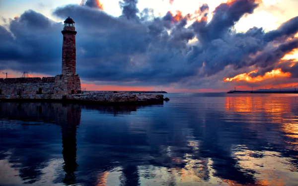 HD desktop wallpaper featuring a brick lighthouse by the sea with a stunning sunset reflecting off the water and dramatic clouds in the sky.