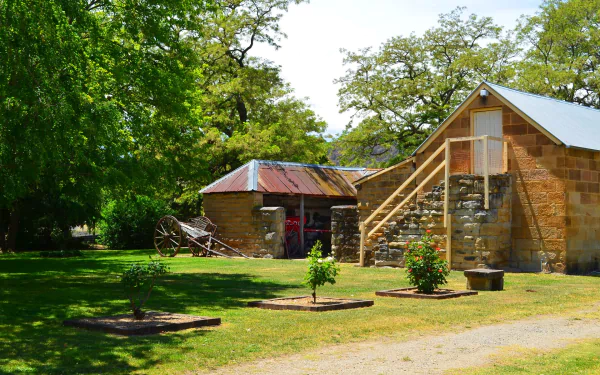 A garden scene at Eskbank House in Lithgow featuring a rustic stone building, an old cart, and lush green trees under clear daylight.