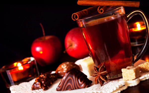 A warm cup of spiced cider garnished with cinnamon sits next to fresh apples, chocolate treats, and flickering candles, creating a cozy Christmas atmosphere in this 4K Ultra HD wallpaper.