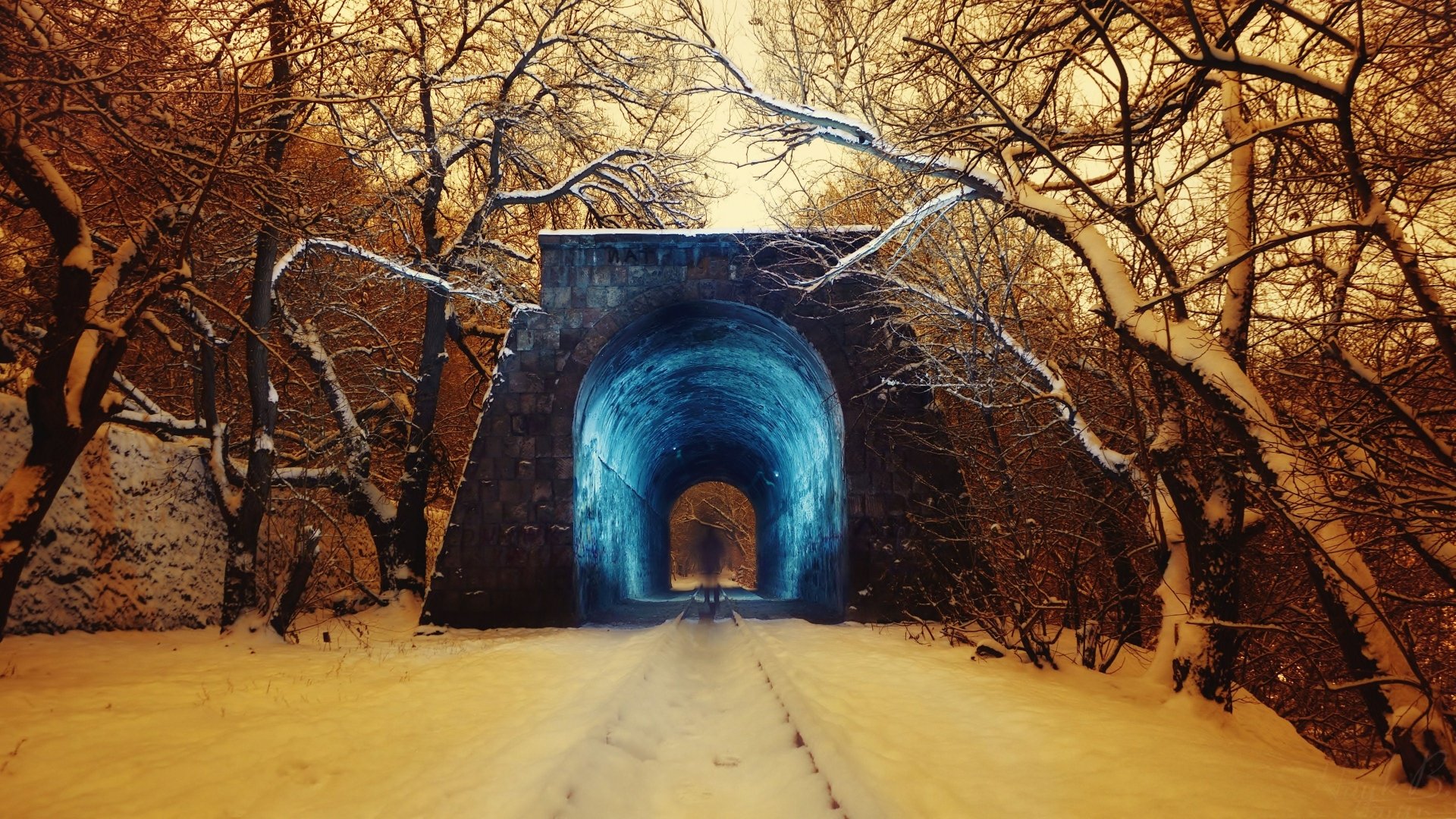 4K Ultra HD winter photography of a snow-covered train track leading through a glowing blue tunnel, framed by bare trees under a golden sky.
