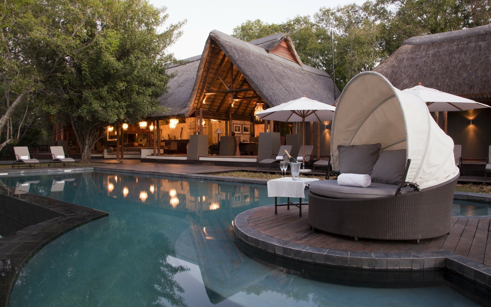 HD PC desktop wallpaper and background — man-made resort pool with circular cabana daybed on a wooden deck, umbrellas and thatched-roof lodge glowing at dusk.