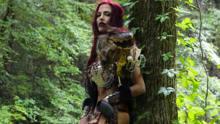 Red Sonja cosplay: woman with long red hair in chainmail armor leaning against a tree in a lush forest — 2K Quad HD PC desktop wallpaper/background