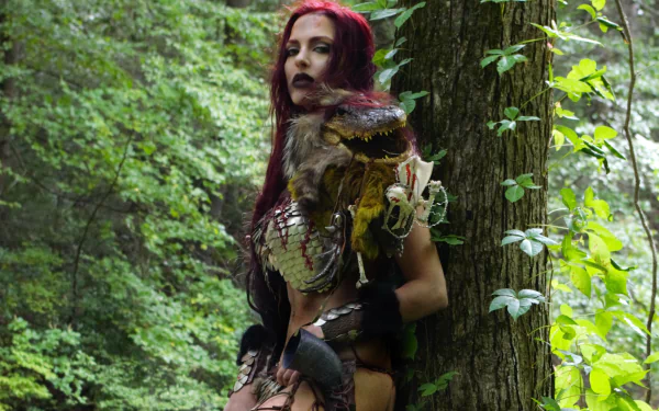 Red Sonja cosplay: woman with long red hair in chainmail armor leaning against a tree in a lush forest — 2K Quad HD PC desktop wallpaper/background