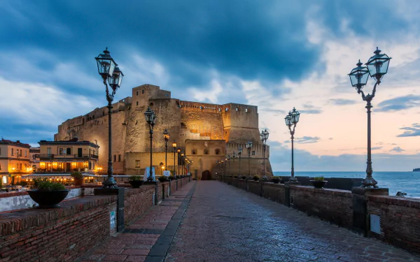 HD PC desktop wallpaper: man-made Castel dell'Ovo at dusk, illuminated seaside fortress and lamp-lit promenade beneath a dramatic cloudy sky.
