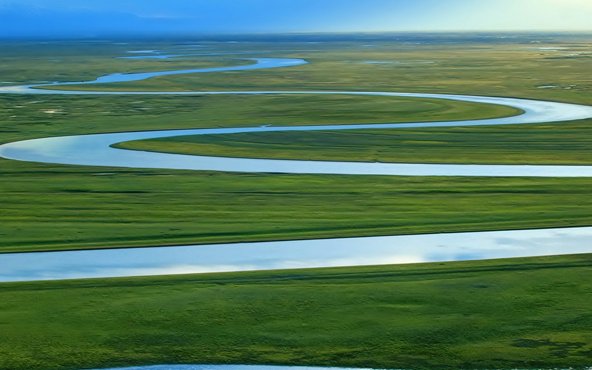 HD PC desktop wallpaper: aerial view of a meandering river cutting through vibrant green plains under a clear blue sky.