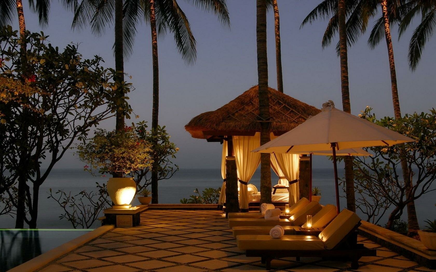 HD PC desktop wallpaper and background of a man-made seaside resort at dusk: lit cabana, lounge chairs and umbrellas on a tiled terrace framed by palm trees overlooking calm ocean.