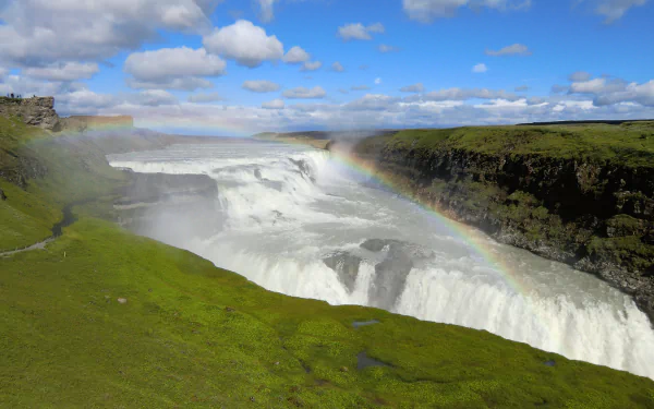 4K Ultra HD desktop wallpaper of Iceland’s Gullfoss waterfall, showcasing cascading water, lush green cliffs, and a vibrant rainbow under a bright blue sky.