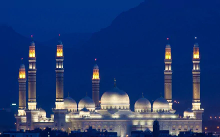 Stunning view of Al Saleh Mosque in Sana'a, Yemen, illuminated at night. The grand architecture features multiple minarets against a dramatic mountain backdrop.