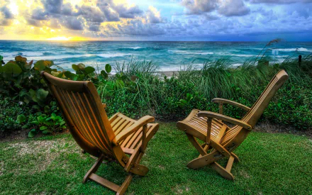 A serene HD desktop wallpaper featuring two wooden chairs overlooking a tranquil beach scene, with waves crashing against the shore and a vibrant sunset in the background.