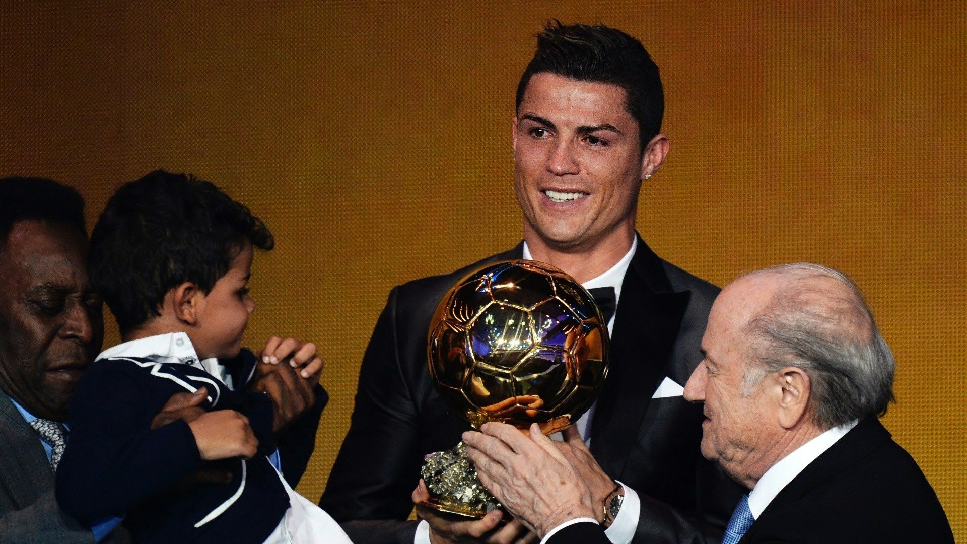 Smiling man in tuxedo holding the Ballon d'Or trophy on stage with officials and a child — Ballon d'Or 2014, Sports HD PC desktop wallpaper background.