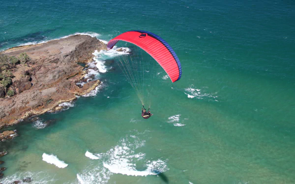 4K Ultra HD desktop wallpaper: red paraglider soars above turquoise sea and rugged coastline, high-energy paragliding sports scene.