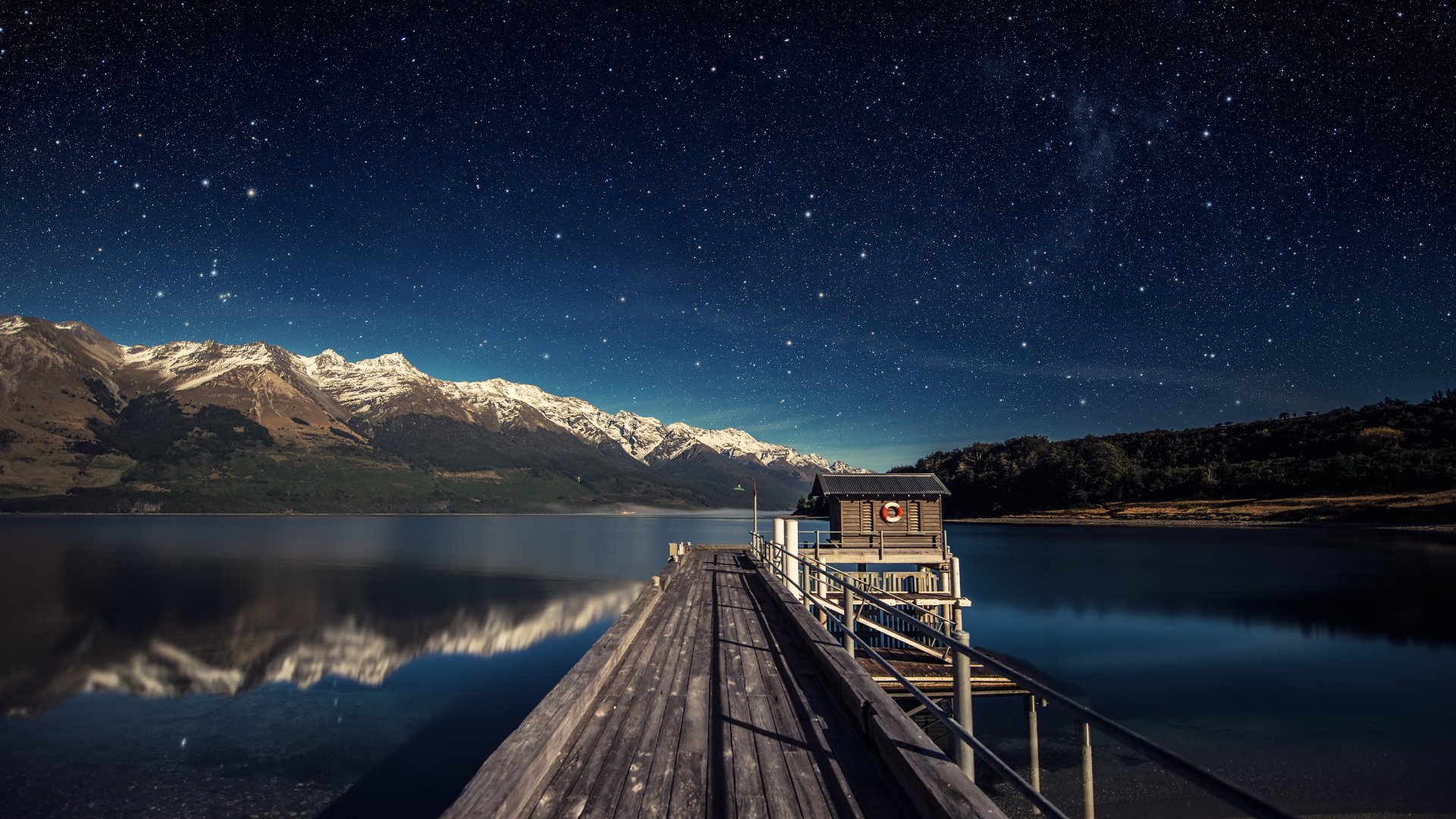 HD desktop wallpaper depicting a starry night sky over a tranquil lake, showing a pier leading to a hut, with the serene reflection of mountains in the background.