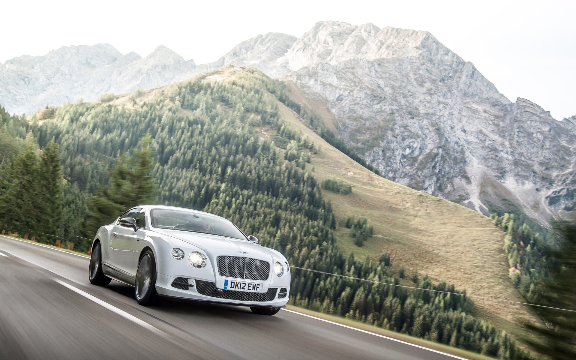 A Bentley Continental GT Speed driving on a mountain road with lush green hills and rocky peaks in the background, captured as an HD PC desktop wallpaper.