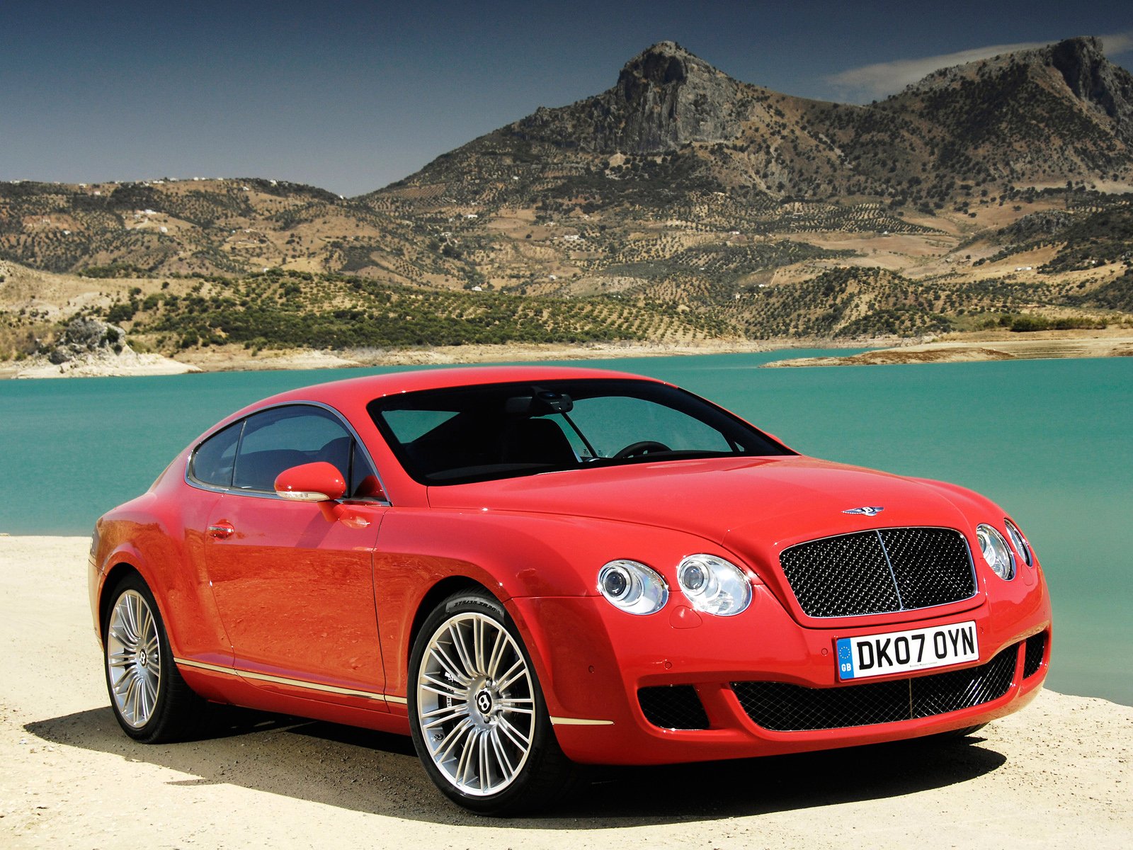 Red Bentley Continental GT Speed parked by a turquoise lakeside with rocky mountains in the background, showcased in an HD PC desktop wallpaper.