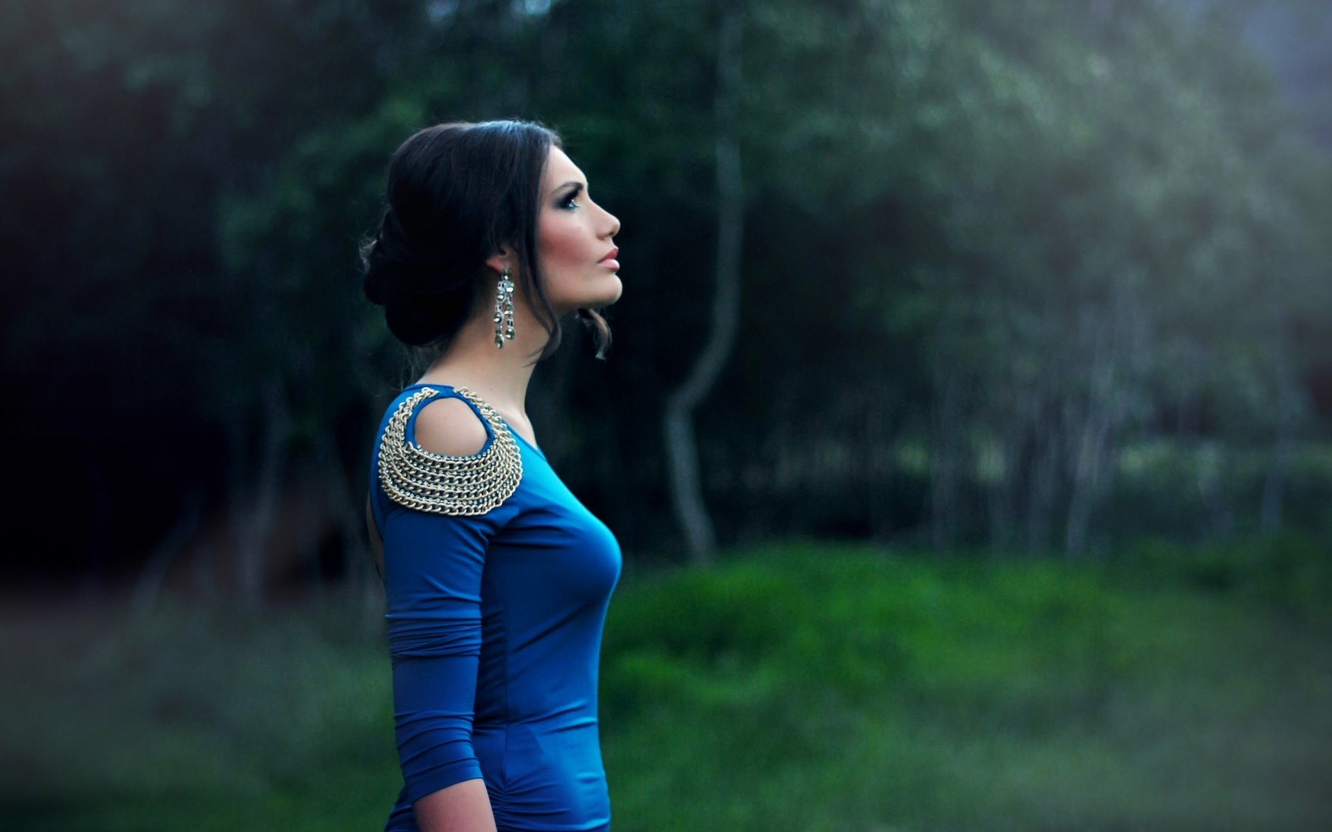 A brunette woman in a blue dress gazes pensively to the side in a lush, forested background. HD desktop wallpaper with a serene, moody atmosphere.