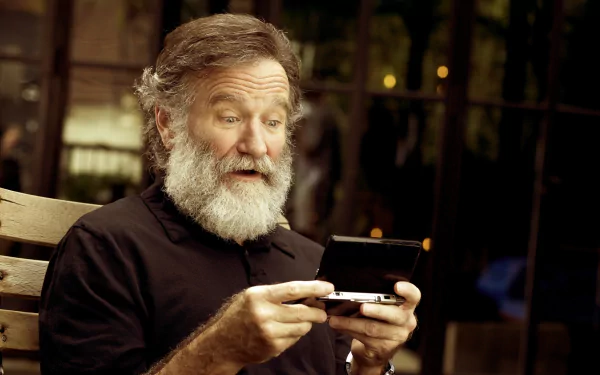 HD desktop wallpaper featuring a person resembling Robin Williams looking at a handheld device with an amused expression while seated on a bench.