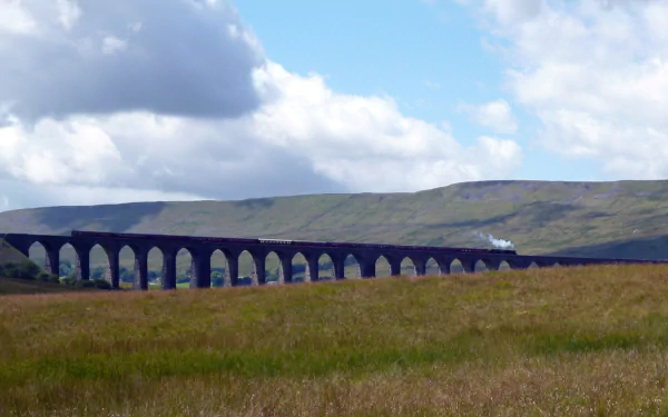 HD desktop wallpaper featuring a steam train crossing a long stone viaduct with grassy fields and rolling hills under a partly cloudy sky.
