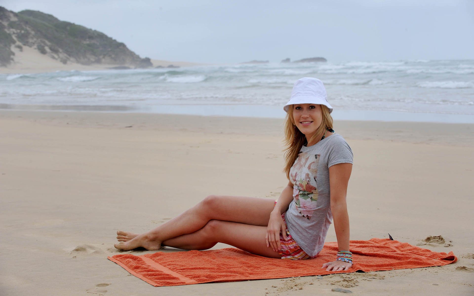 German actress Wolke Hegenbarth sits smiling on an orange towel at a serene beach, featured in an HD PC desktop wallpaper and background.