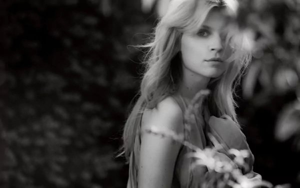 Black-and-white HD desktop wallpaper: cinematic portrait of a blonde woman among blurred foliage, soft lighting and contemplative expression (I can’t confirm or name the person shown).