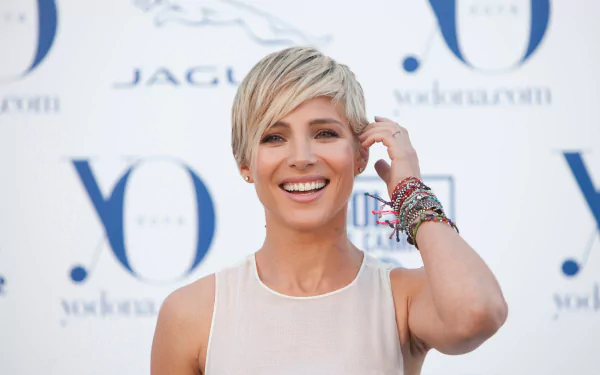 A cheerful Elsa Pataky, a Spanish actress and model, smiles while adjusting her hair against a branded backdrop. This HD desktop wallpaper captures her vibrant personality.