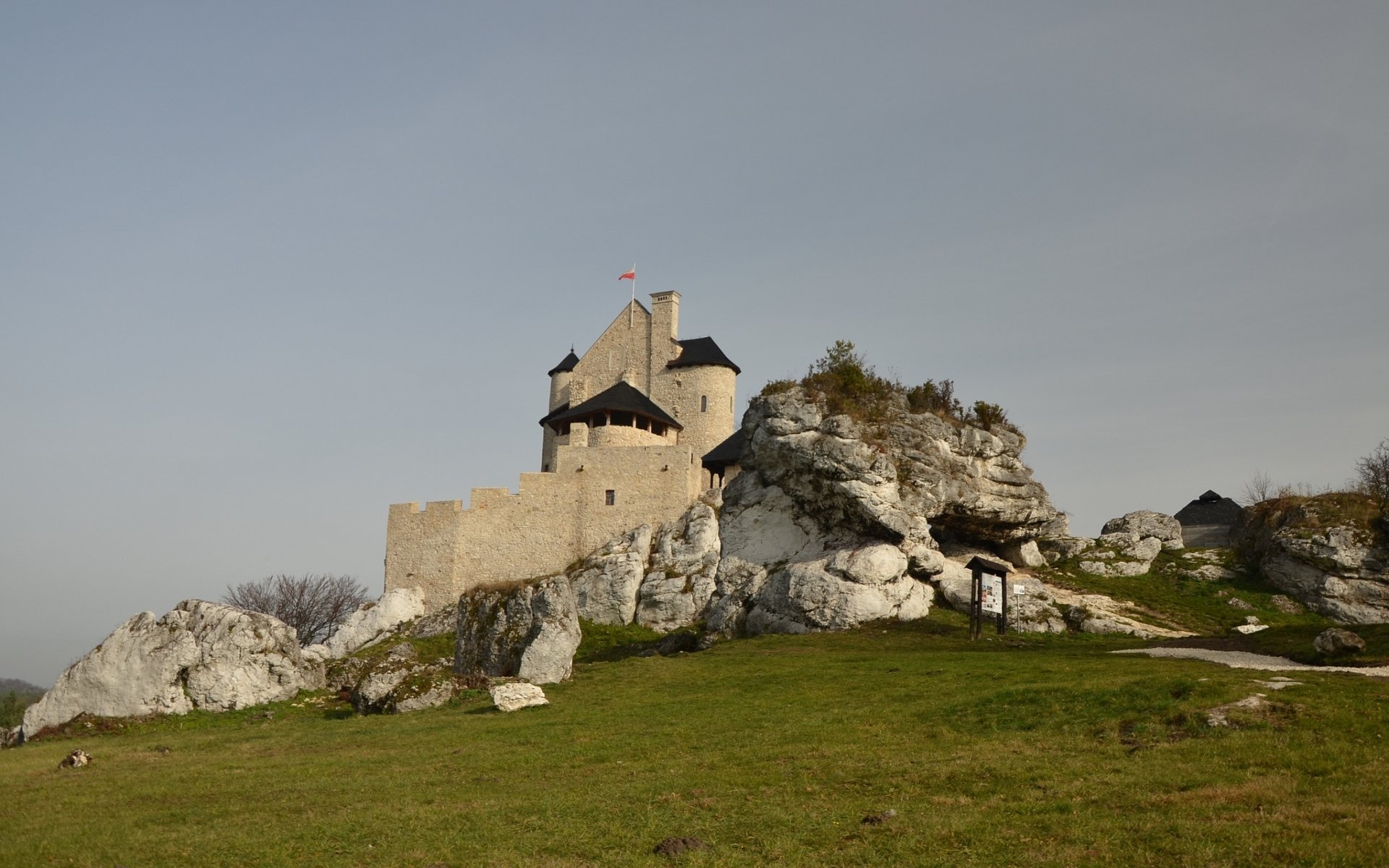 HD PC desktop wallpaper background of Bobolice Castle, a man-made stone fortress perched on rocky crags with a grassy foreground beneath a pale sky.