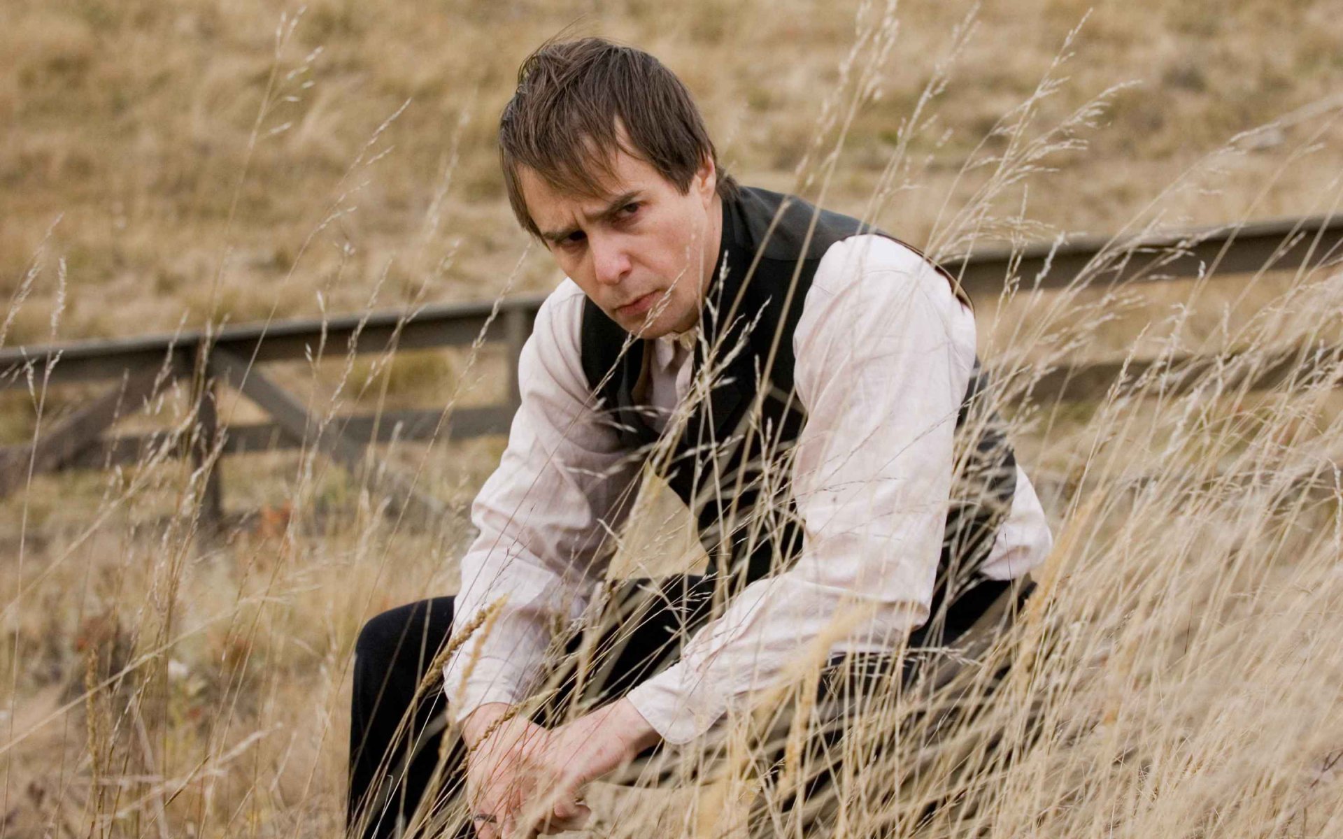 2K Quad HD PC desktop wallpaper — brooding man in period vest crouches in tall grass, scene from The Assassination of Jesse James by the Coward Robert Ford.