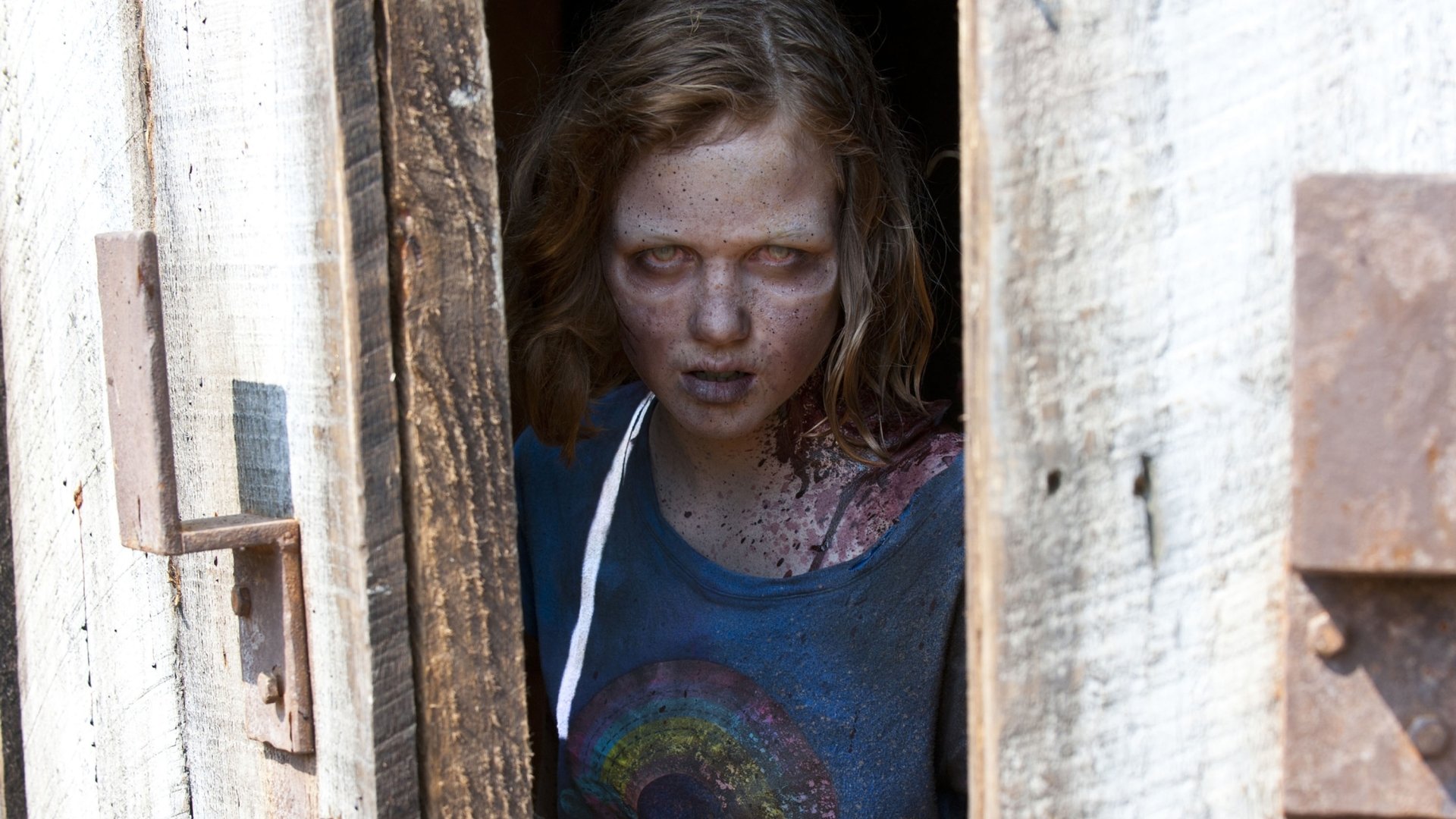 A chilling moment from the TV show The Walking Dead, featuring a disheveled girl with a haunting gaze, peering through a wooden door, capturing the eerie essence of the series.