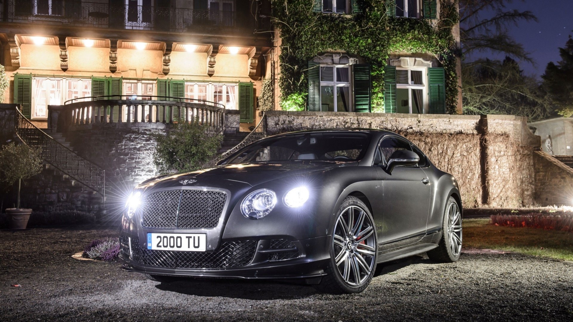 HD PC desktop wallpaper and background: matte-black Bentley Continental GT Speed parked at night before an illuminated manor, headlights on.