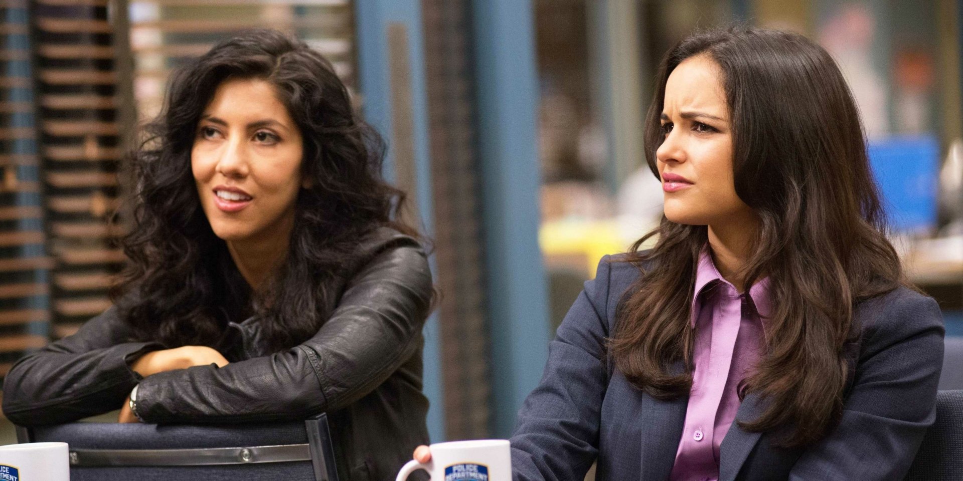 Two characters from the TV show Brooklyn Nine-Nine, featuring Stephanie Beatriz, are seated in a bustling precinct environment, showcasing their distinct personalities and styles.