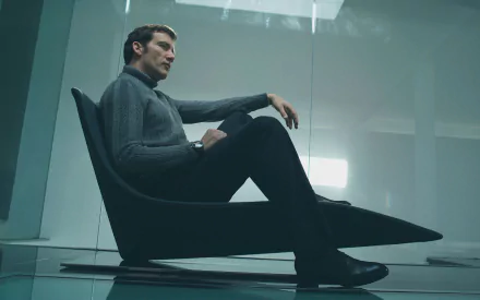 Cinematic 2K Quad HD PC desktop wallpaper: a man in a dark turtleneck reclines in a low modern chair inside a minimalist glass-walled room, moody lighting and reflective floor.