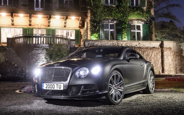 HD PC desktop wallpaper and background: matte-black Bentley Continental GT Speed parked at night before an illuminated manor, headlights on.