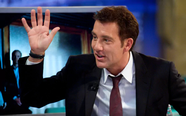 Man in a dark suit and burgundy tie raising his hand while speaking on a TV talk-show set — 2K Quad HD PC desktop wallpaper/background.