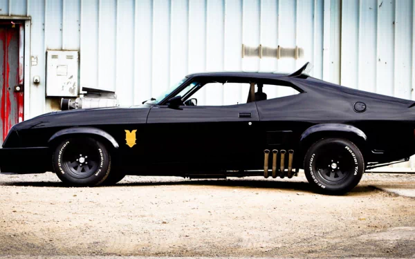 HD PC desktop wallpaper showing a side profile of a black Mad Max Interceptor muscle car parked against a corrugated metal wall.