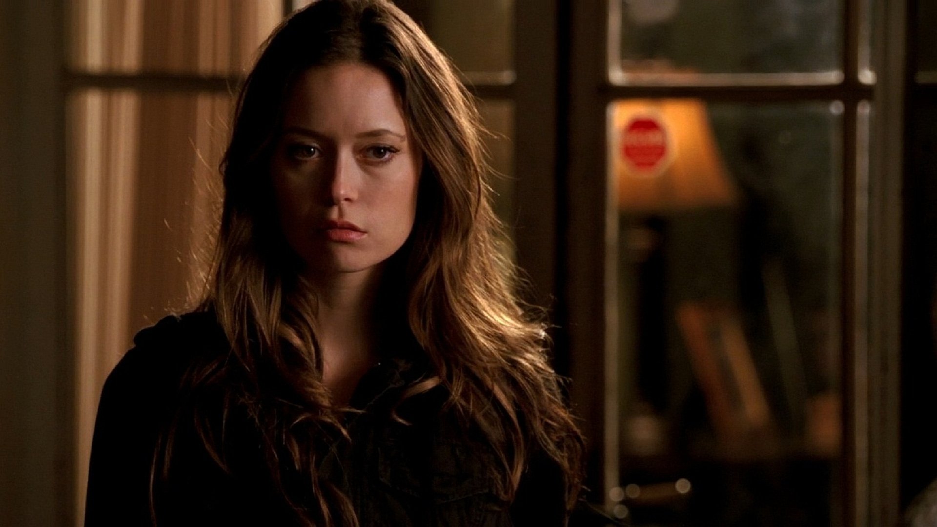 HD PC desktop wallpaper/background of a celebrity actress with long brown hair and a serious expression, standing in a dim room with a lamp and a windowed door behind her.