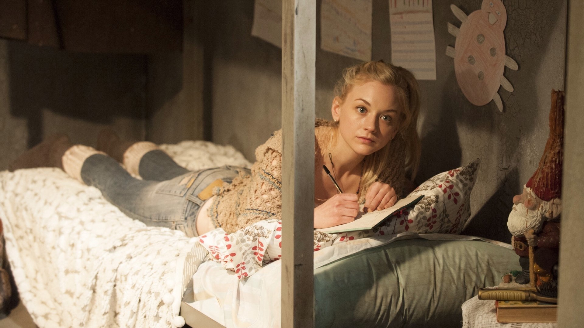 A character from The Walking Dead lies on a bed, writing in a notebook in a dimly lit room decorated with drawings, captured in this HD PC desktop wallpaper.