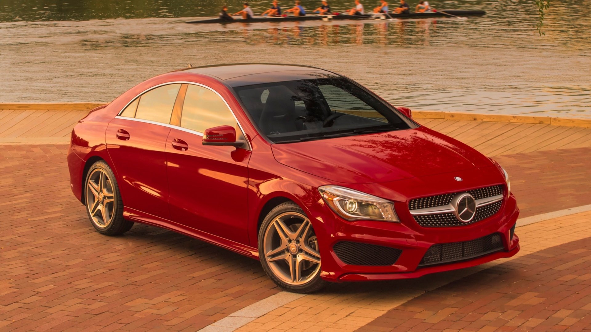 HD PC desktop wallpaper and background showing a red Mercedes‑Benz CLA‑Class vehicle parked on a riverside promenade, with rowers on the water in the distance.