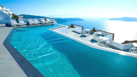A stunning view of a resort pool in Santorini, Greece, overlooking the ocean, surrounded by lounge chairs and a serene landscape of blue waters and sky.