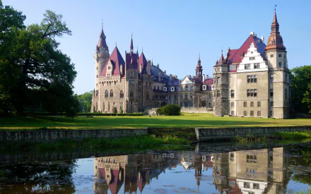 HD PC desktop wallpaper: Moszna Castle, a man-made chateau with red roofs and turrets reflected in a calm moat beneath a clear blue sky.