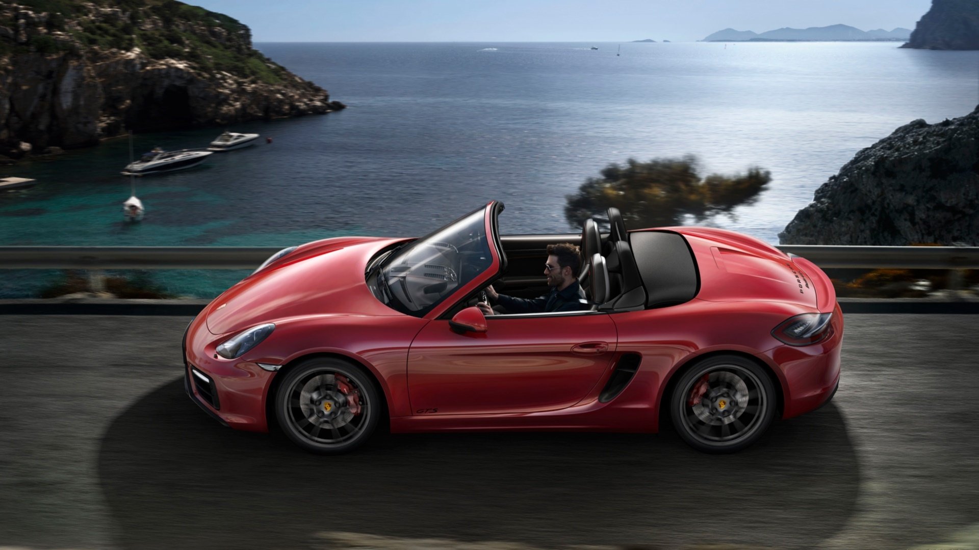Red Porsche Boxster GTS convertible cruising a coastal road with rocky cliffs and calm sea — HD PC desktop wallpaper background.